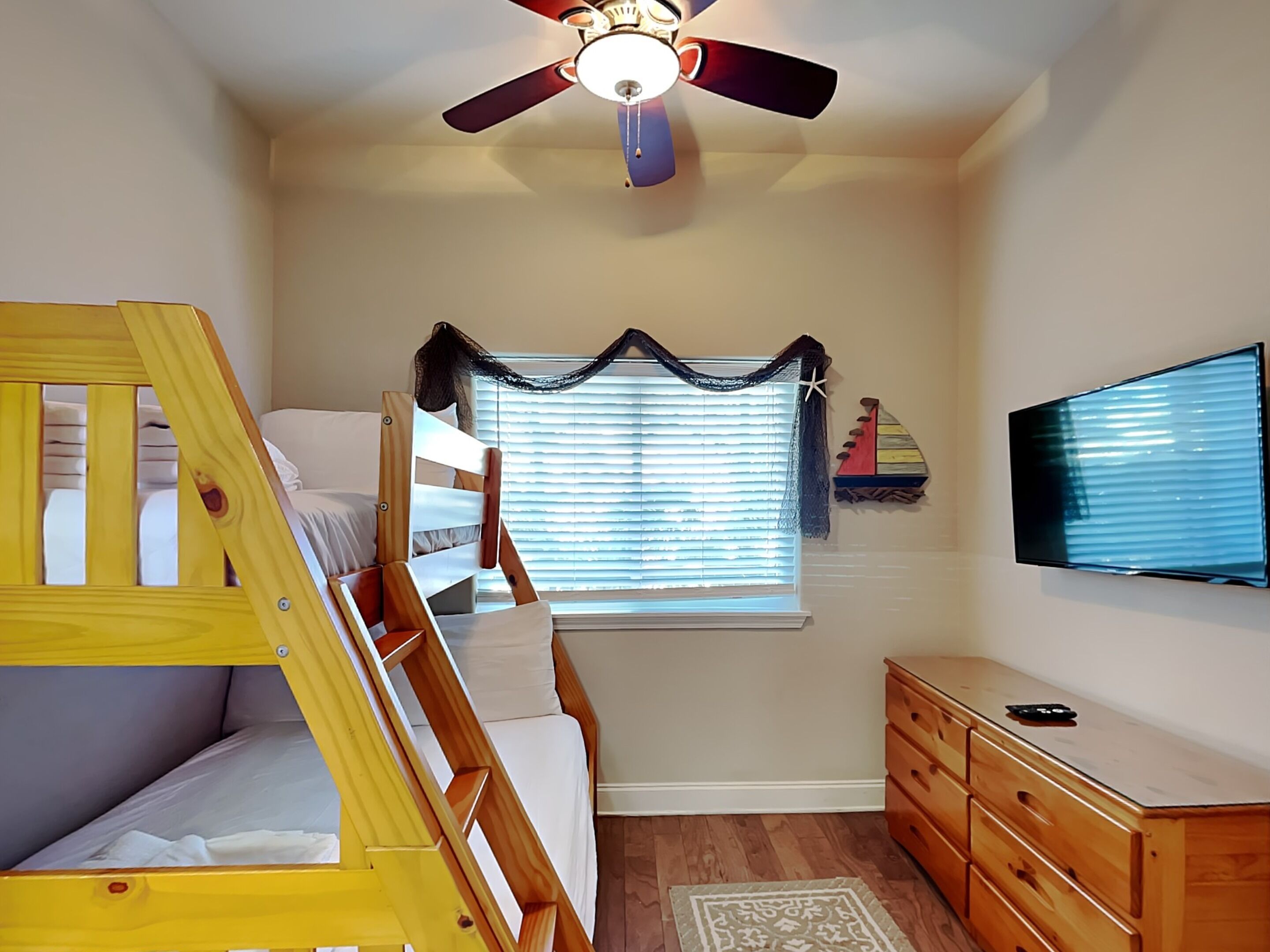 Quarto com cama beliche, ventilador de teto, janela com persianas, televisão e cômoda.