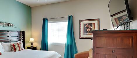 A bedroom with a bed, a nightstand, a desk, a chair, a ceiling fan, a window with curtains, and a framed picture on the wall.