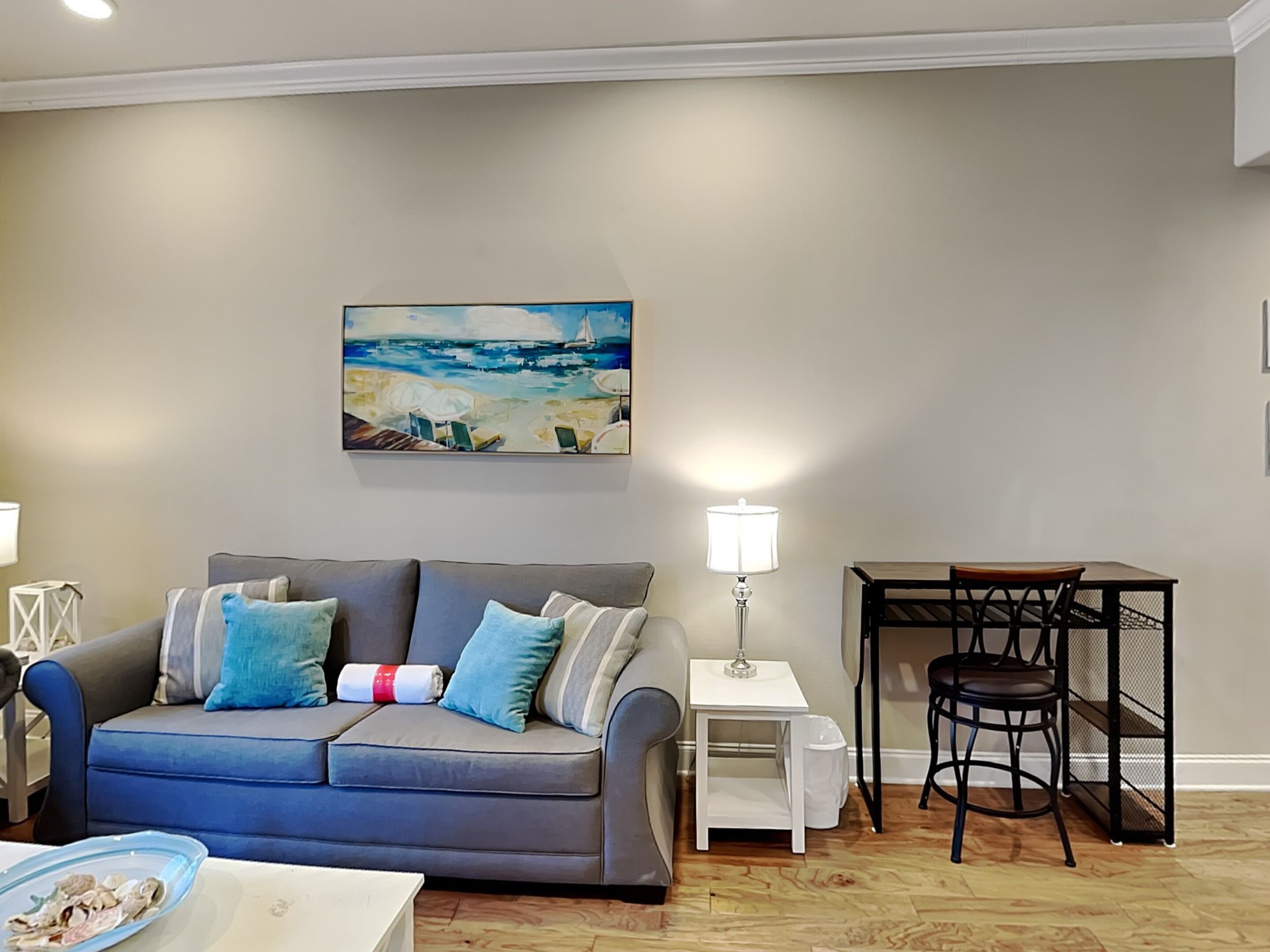 Un salon avec un canapé gris, une petite table, une lampe et un tableau représentant une scène de plage.