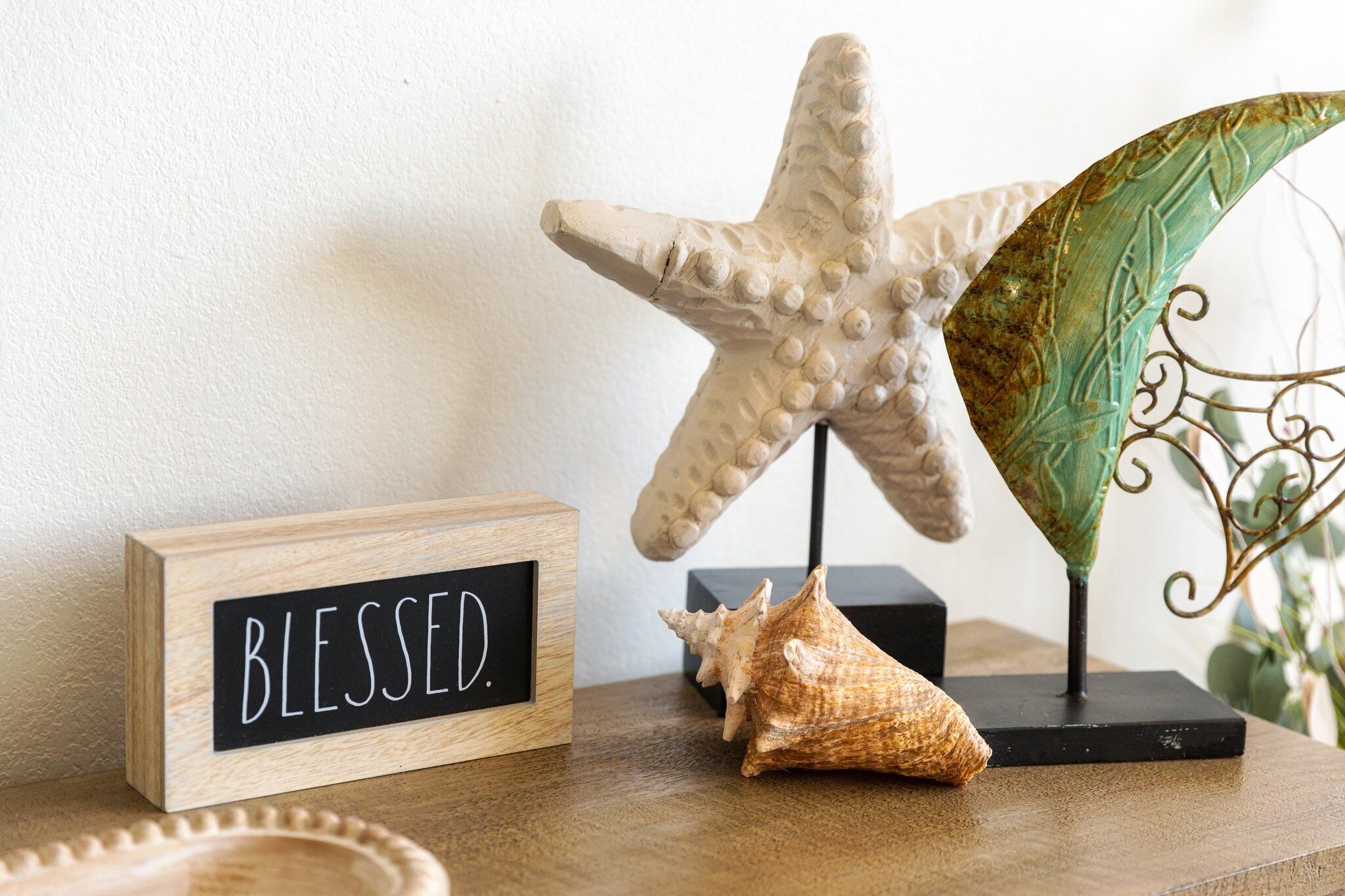 A wooden block with "BLESSED." written on it, a starfish, and a seashell on a wooden surface.