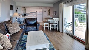 Un salon avec un canapé en cuir marron, une table basse blanche et un coin repas avec une table et des chaises blanches. On aperçoit une cuisine au fond.