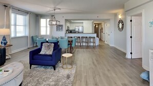 A spacious living room with a blue sofa, a round coffee table, and a ceiling fan. A kitchen with white cabinets and a bar area is visible in the background. - Ariel Dunes II by Southern Vacation Rentals (Miramar Beach)