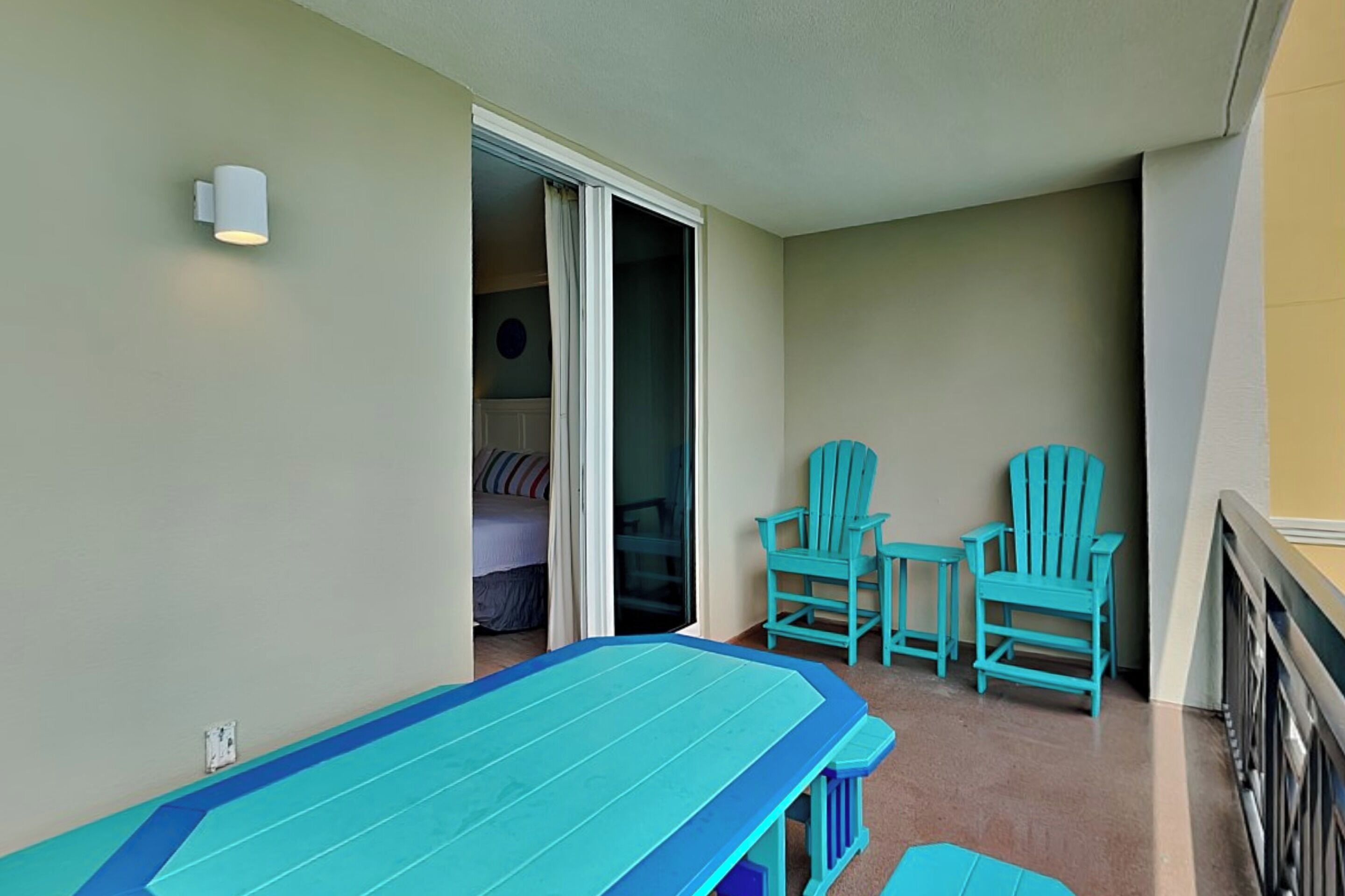A balcony with a blue table and two turquoise chairs, a sliding glass door leading to a bedroom, and a white wall with a light fixture.