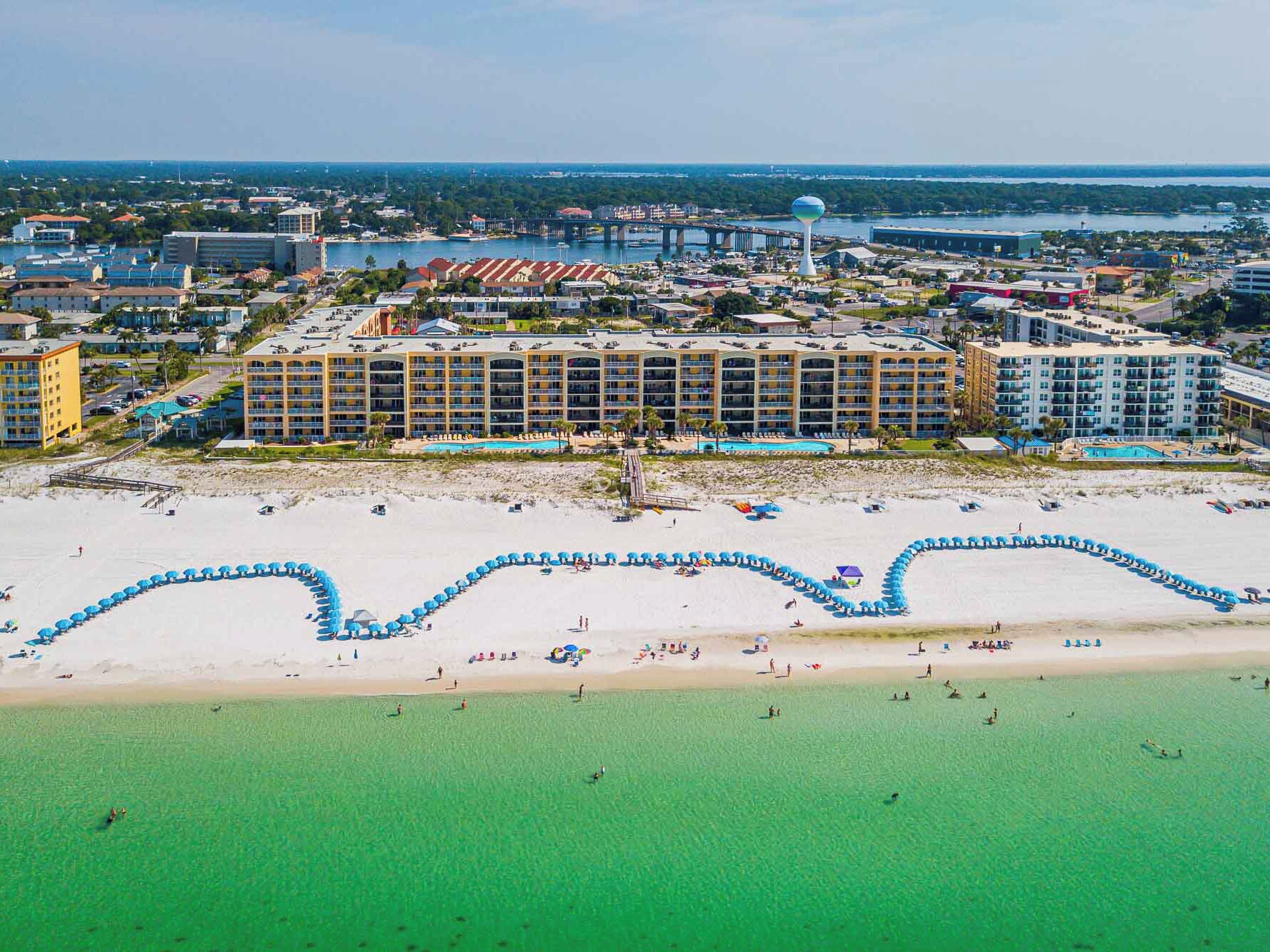 A coastal hotel with multiple buildings, a beach with umbrellas, and a clear view of the ocean.