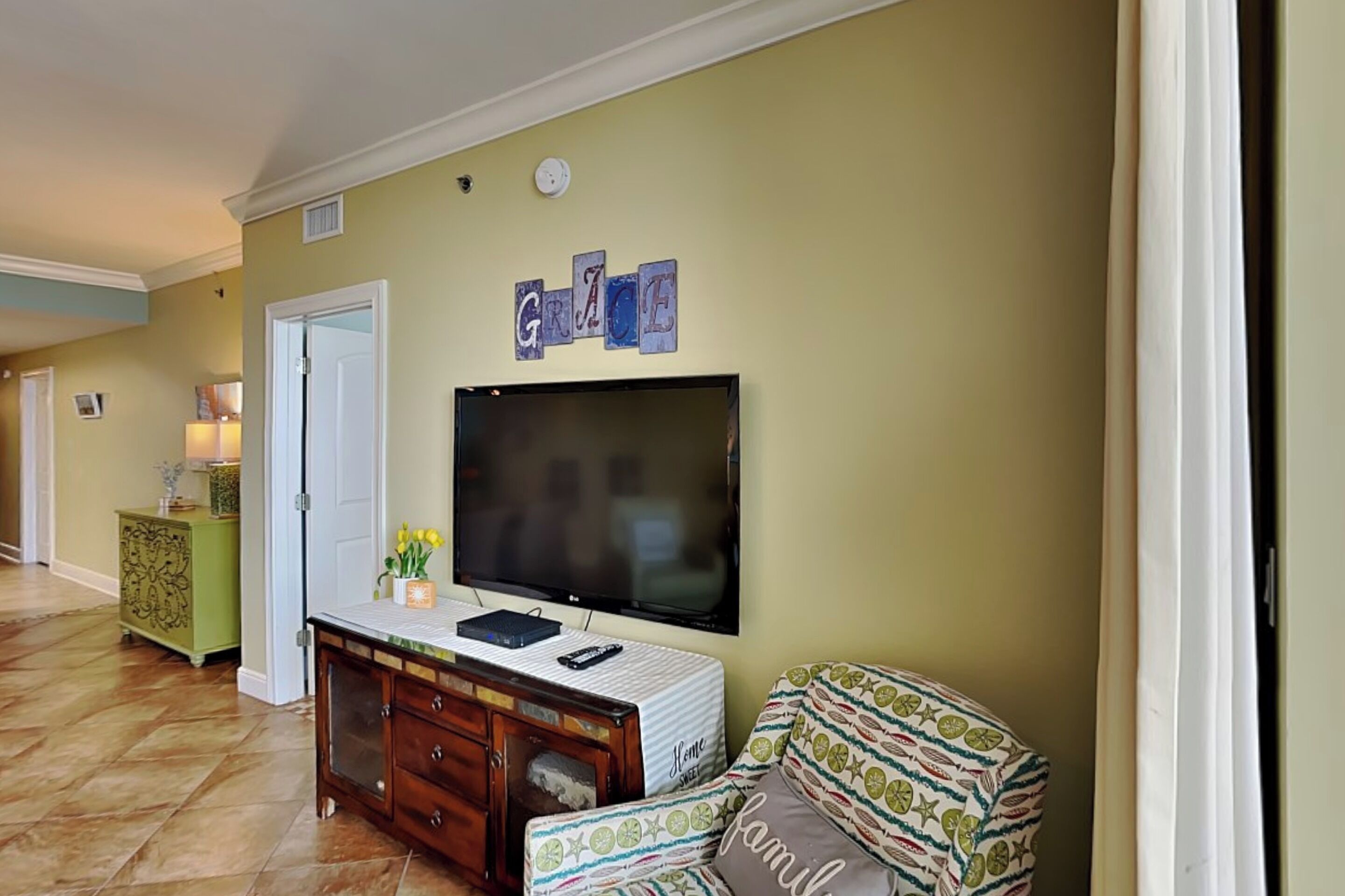 Una sala de estar con un televisor de pantalla plana montado en la pared, un sofá con cojines estampados, un aparador de madera y una mesita auxiliar verde.