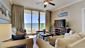 A living room with a sofa, armchair, and coffee table, a TV, and a balcony with a view of the ocean.