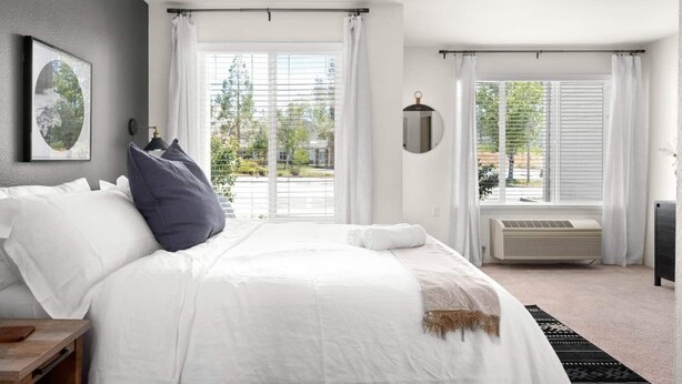Une chambre moderne avec un grand lit, une table de chevet, un climatiseur fixé au mur et une fenêtre donnant sur l’extérieur.
