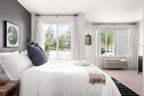 Un dormitorio moderno con una cama grande, una mesita de noche, aire acondicionado de pared y una ventana con vistas al exterior.