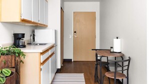 Une petite cuisine équipée d’une machine à café, d’un évier et d’un meuble en bois. Une porte donne accès à une autre pièce, et on y trouve une petite table avec une chaise.
