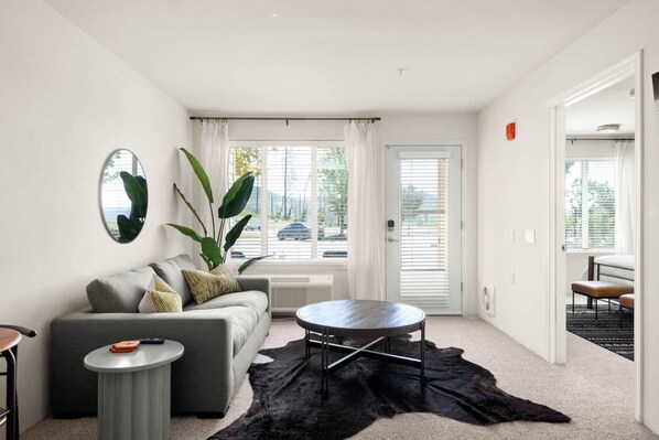 A modern living room with a grey sofa, a round coffee table, and a large black rug. There is a large window with blinds, a door, and a view of the street outside.