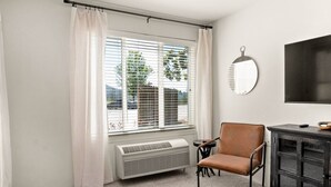A room with a large window, a chair, a small table, a heater, and a mirror on the wall. - The Trinity | Studio in Paradise (Paradise)