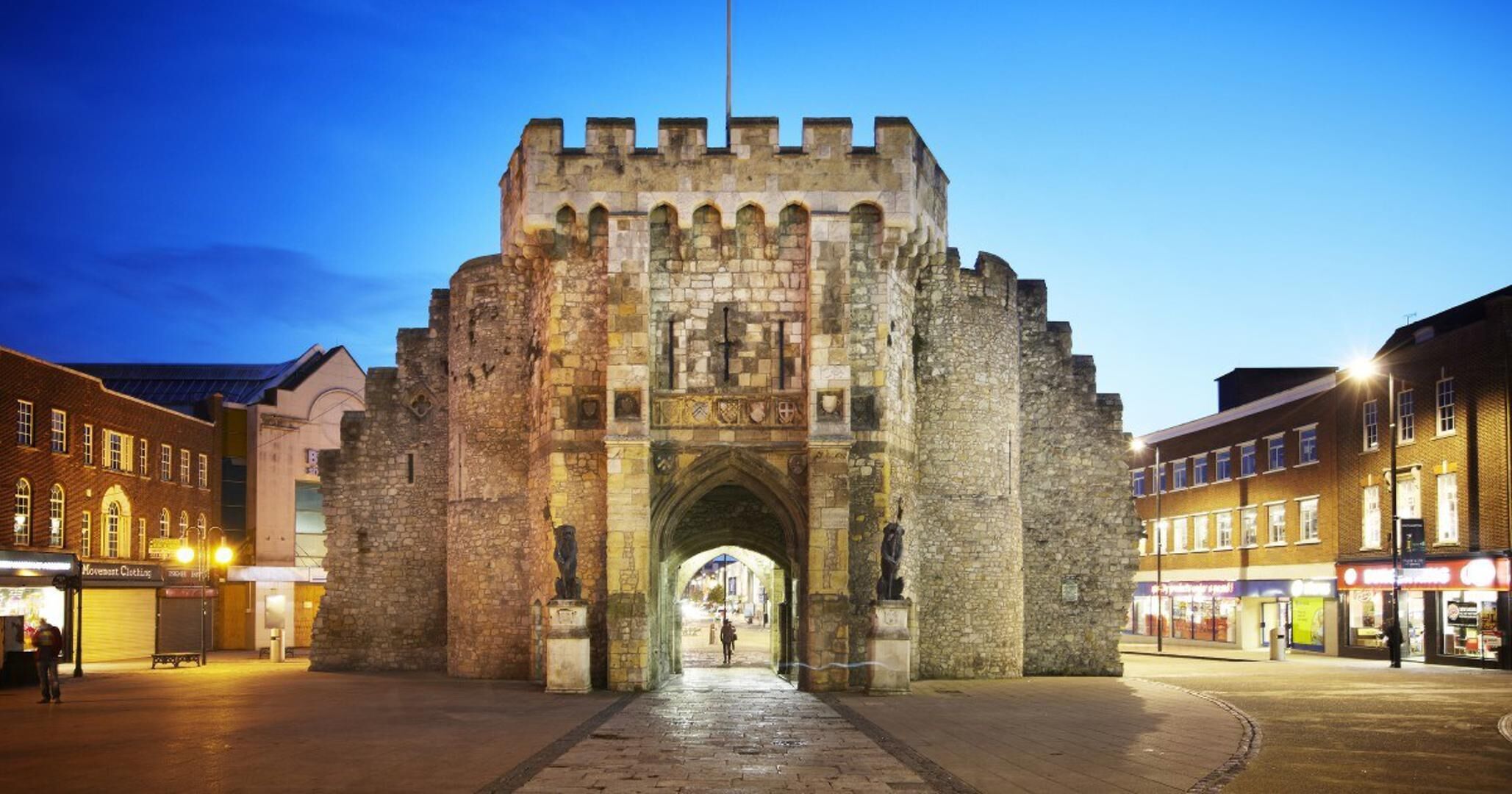 A historic stone archway with intricate carvings and a flagpole, leading into a town square with modern buildings and shops.