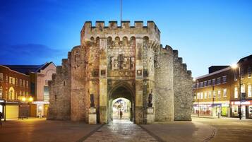 A historic stone archway with intricate carvings and a flagpole, leading into a town square with modern buildings and shops.