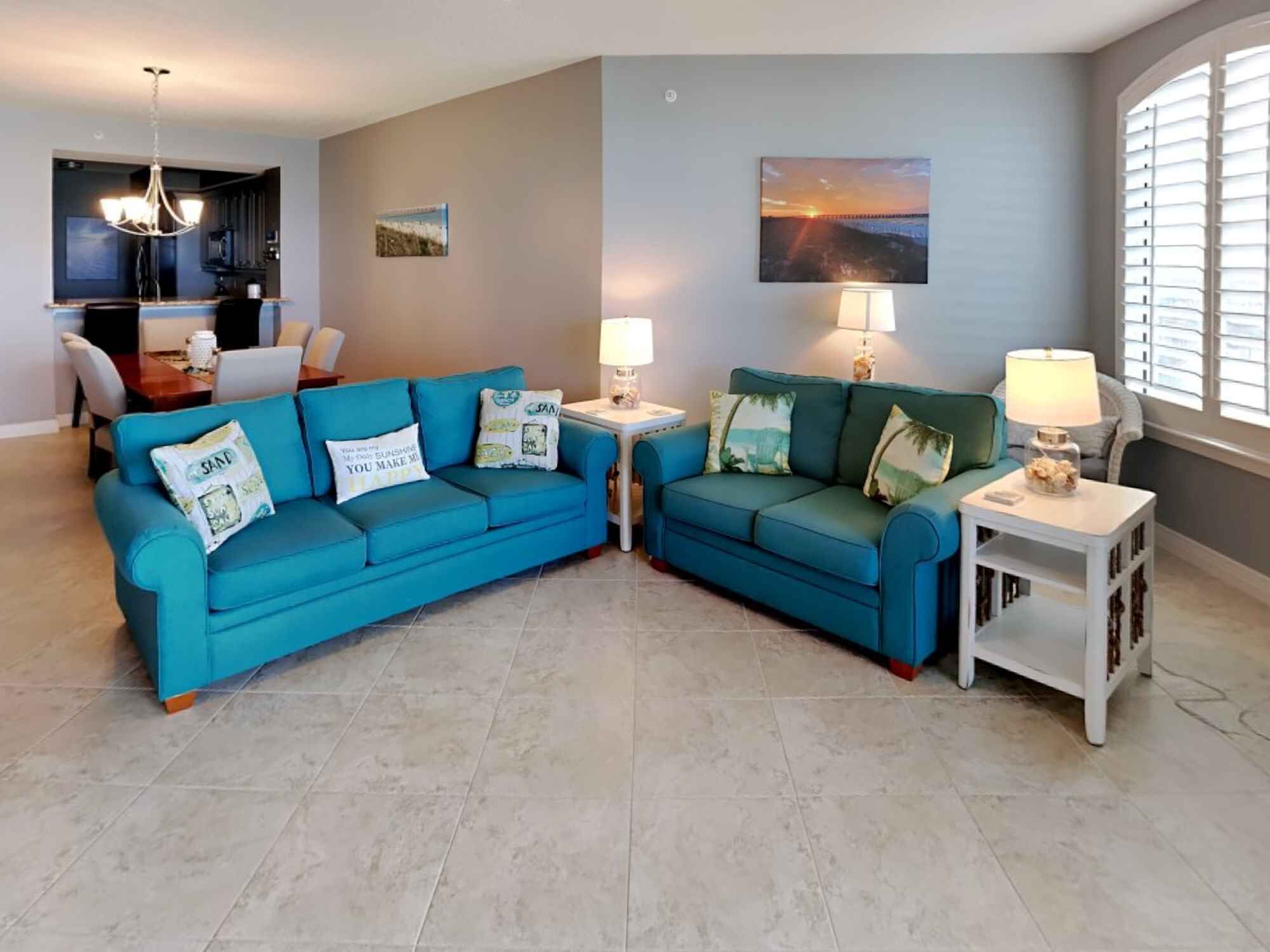 Ein Wohnzimmer mit zwei blauen Sofas, einem Couchtisch und einer Lampe. Im Hintergrund ist ein Essbereich mit einem Kronleuchter zu sehen.