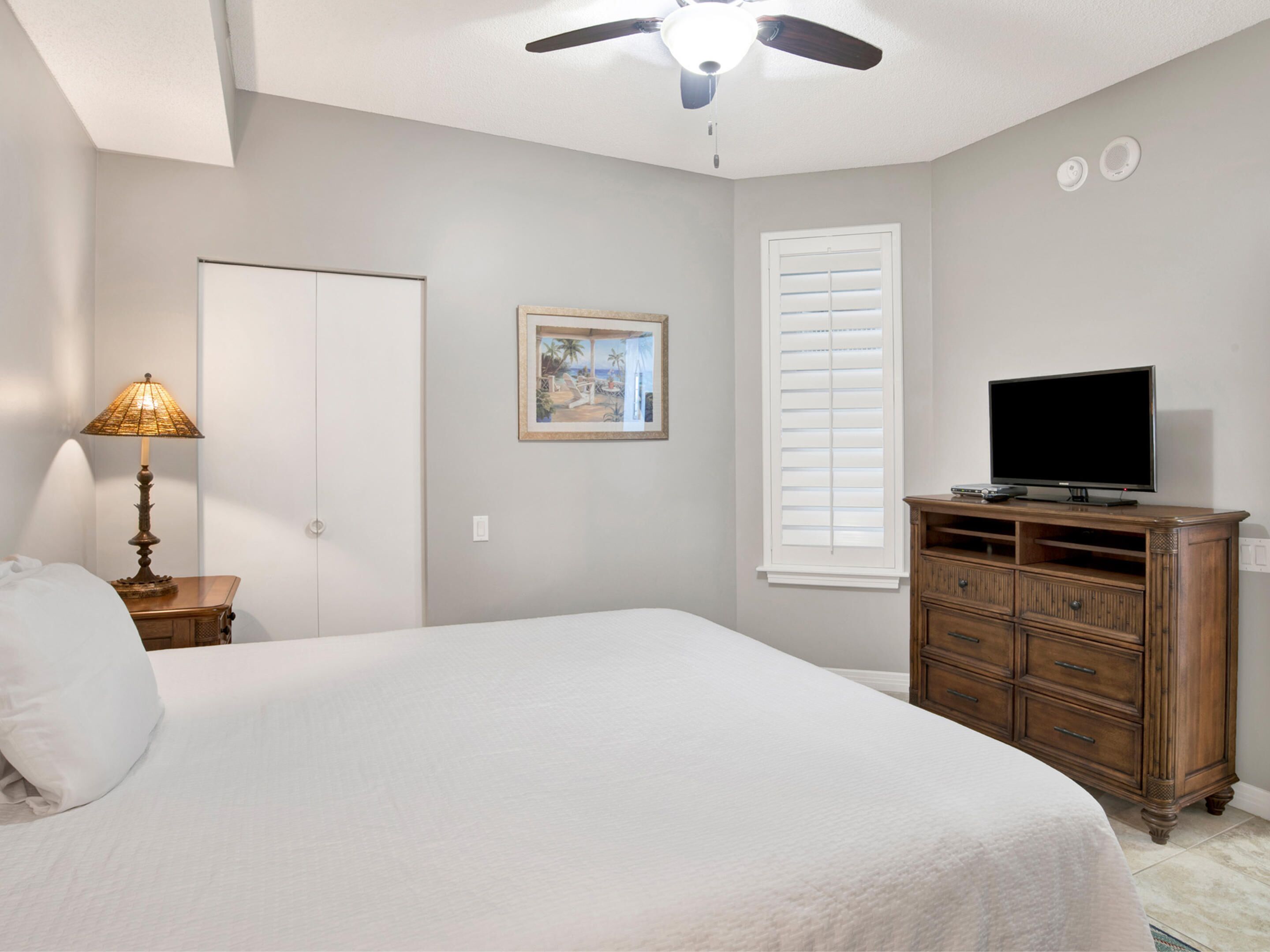 A bedroom with a bed, a dresser, a television, a ceiling fan, a window with blinds, and a door.