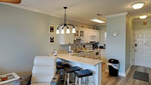 A modern kitchen with white cabinets, a central island with bar stools, and a dining area with a white sofa. - Baywatch by Southern Vacation Rentals (Pensacola Beach)