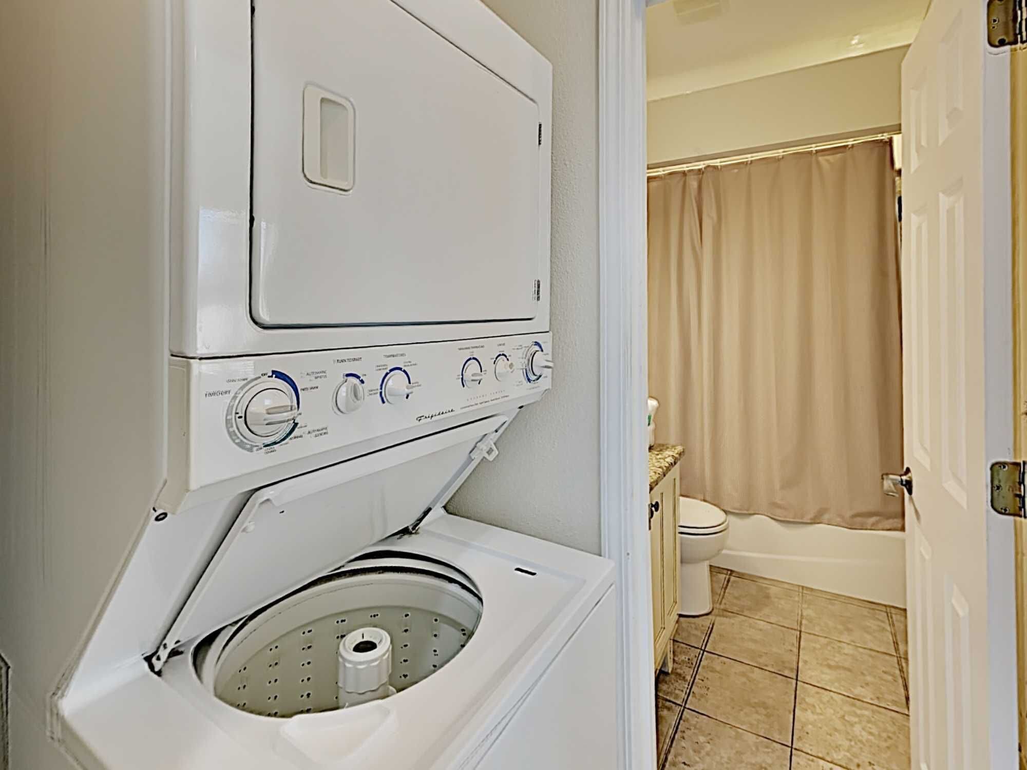 A front-loading washer and dryer unit in a bathroom with a toilet and shower curtain.