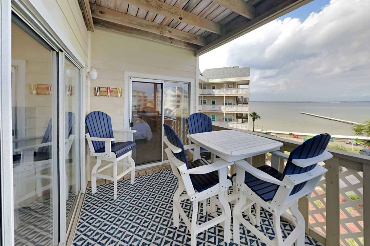 A balcony with a white table and four Adirondack chairs, overlooking a body of water and a building with multiple balconies.