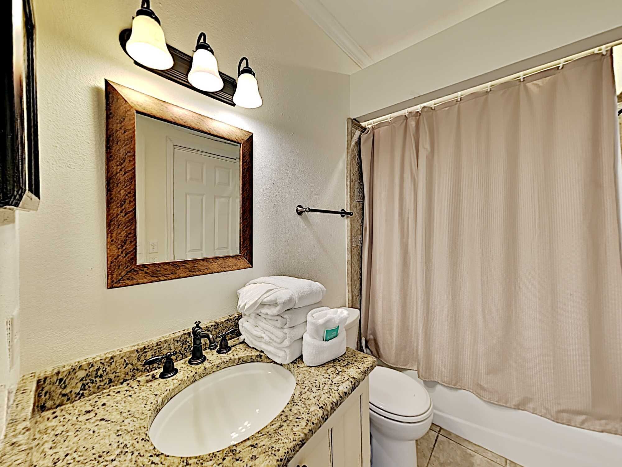 A bathroom with a granite countertop sink, a mirror with a wooden frame, a towel rack with folded towels, a toilet, and a beige shower curtain.