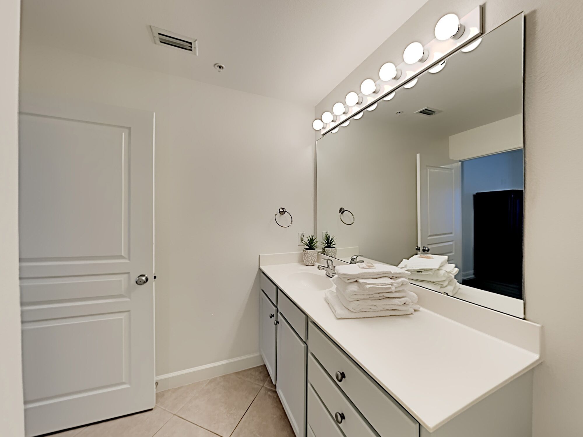 A modern bathroom with a vanity, a large mirror with lights, a towel rack, and a door.