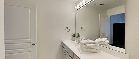 A modern bathroom with a vanity, a large mirror with lights, a towel rack, and a door.