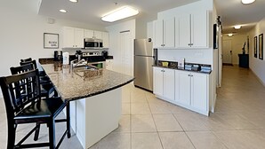 Cozinha moderna com armários brancos, bancada de granito, eletrodomésticos de aço inoxidável e um espaço de jantar com cadeiras pretas.