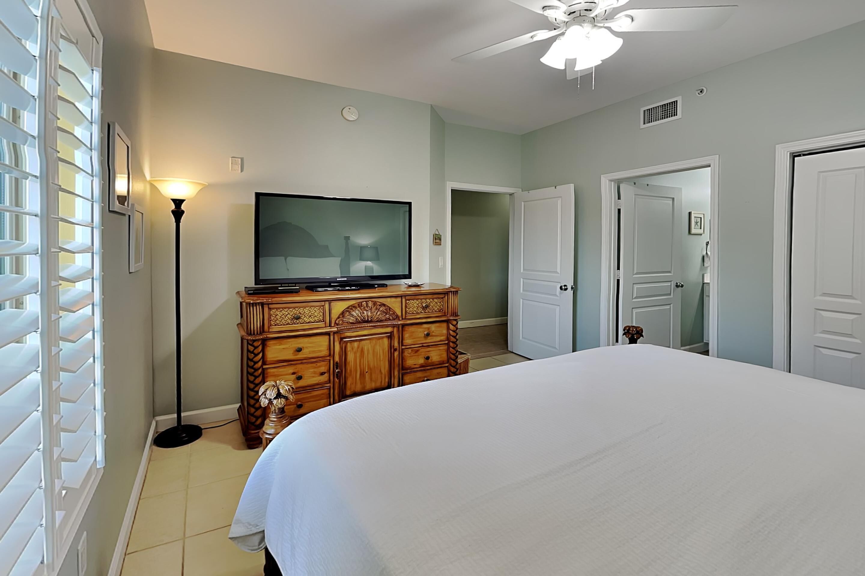 A bedroom with a large bed, a wooden dresser, a television, and a ceiling fan.