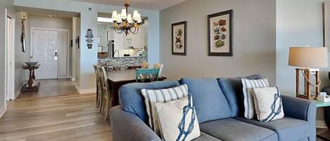 A living room with a blue sofa, a chandelier, framed artwork, and a dining area in the background.