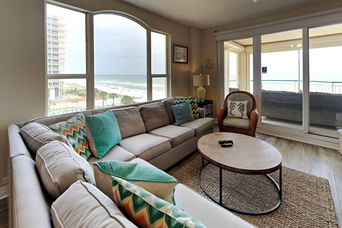 A modern living room with a large sofa, a round coffee table, and a chair. There are large windows offering a view of the beach and ocean.