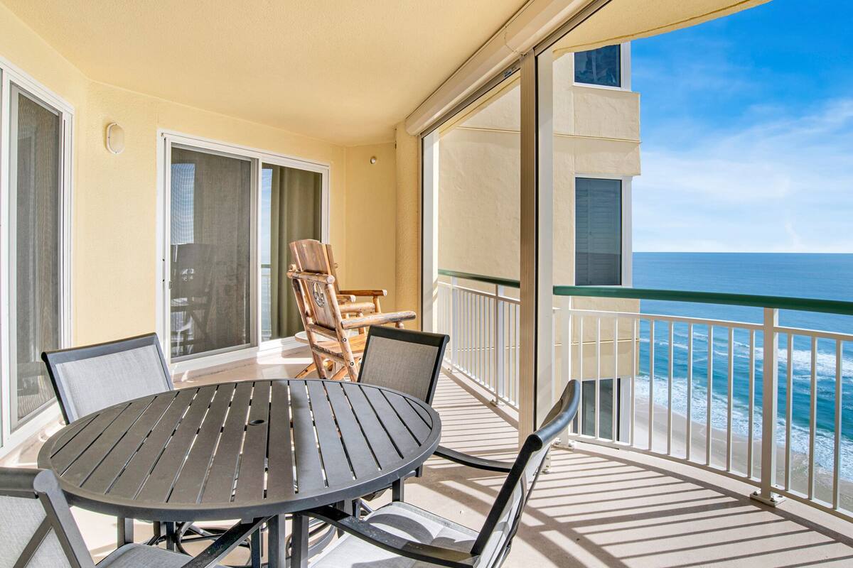 A balcony with a round table and chairs, a view of the ocean, and a building in the background.