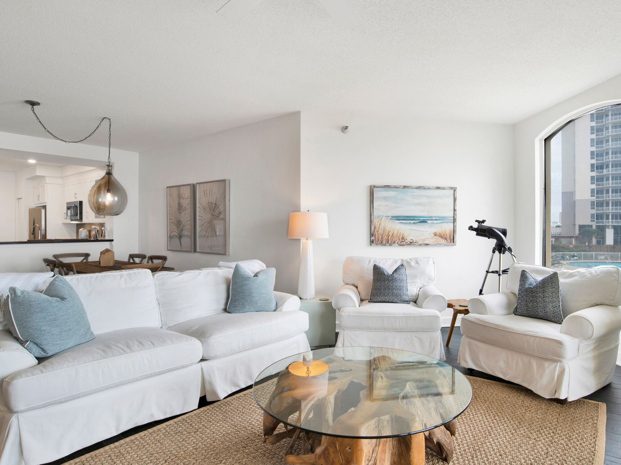 A modern living room with a white sofa set, a glass coffee table, and a beach-themed painting.
