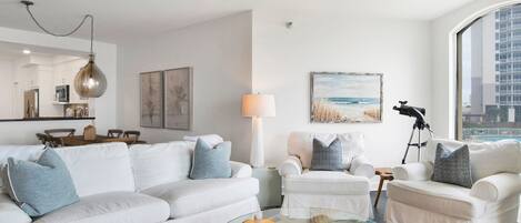 A modern living room with a white sofa set, a glass coffee table, and a beach-themed painting.