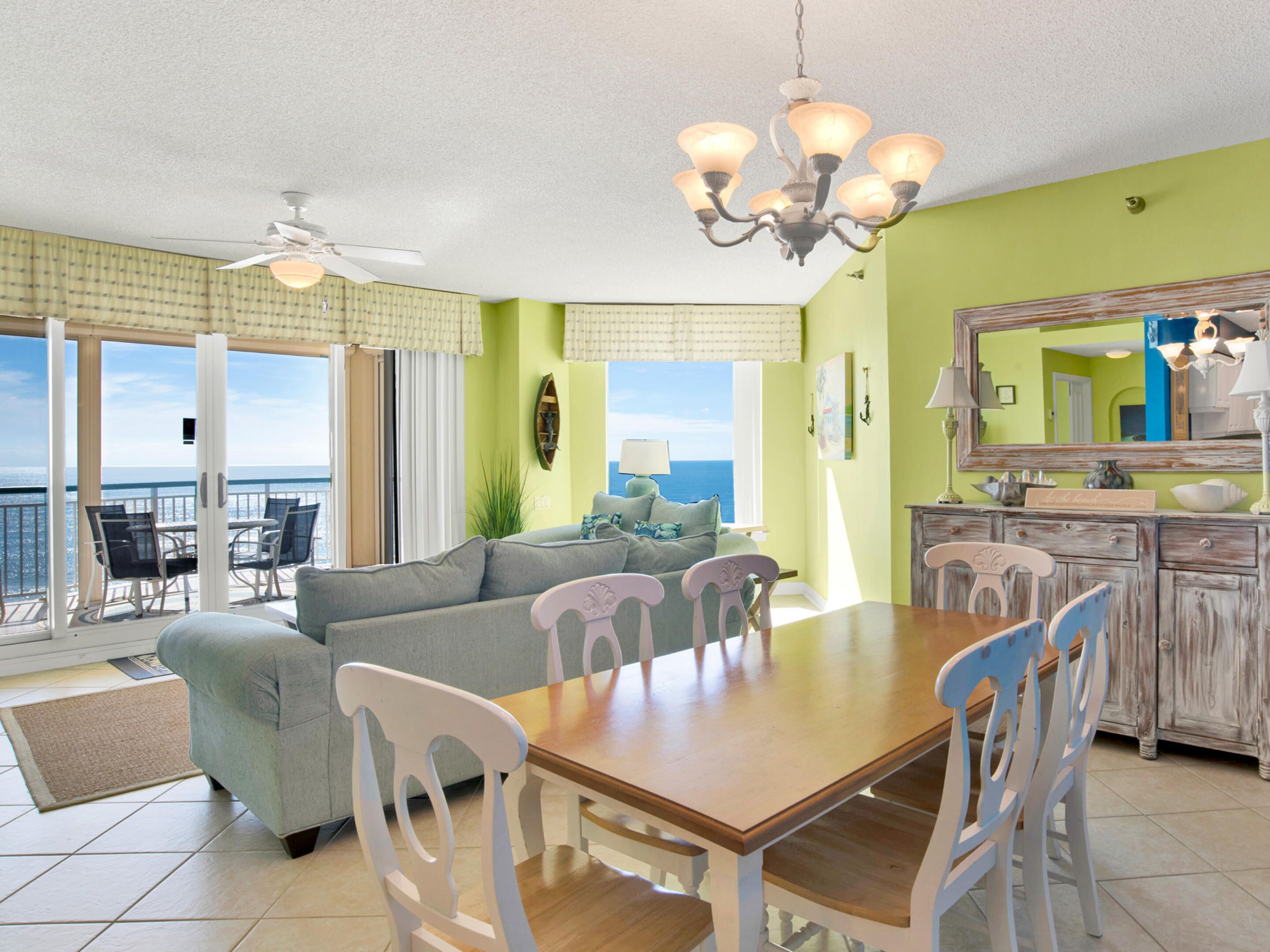 A dining area with a wooden table and white chairs, a ceiling fan, and a view of the ocean through large windows.