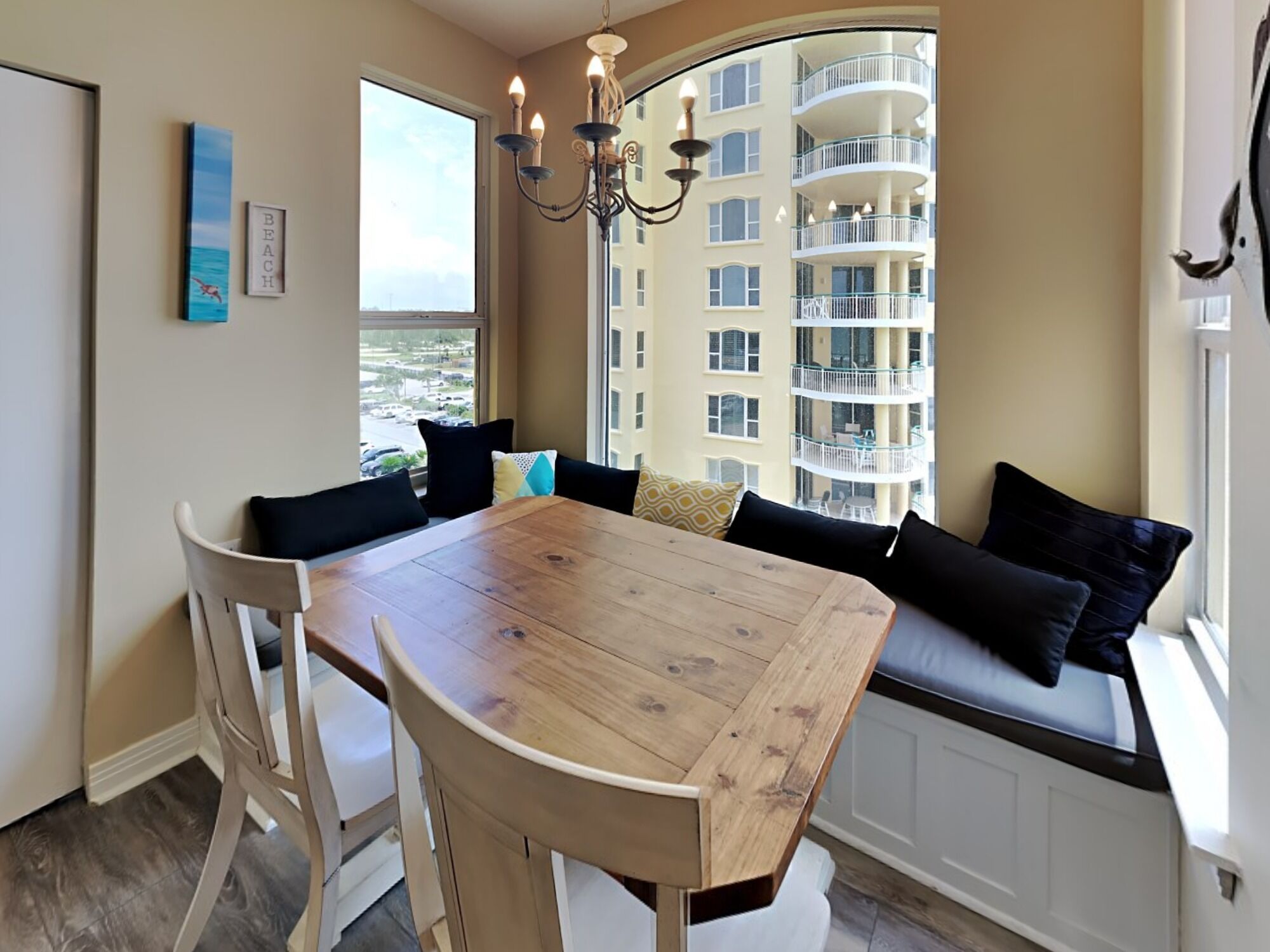 A dining area with a wooden table and white chairs, a bay window with a view of a building and the sea, and a chandelier.