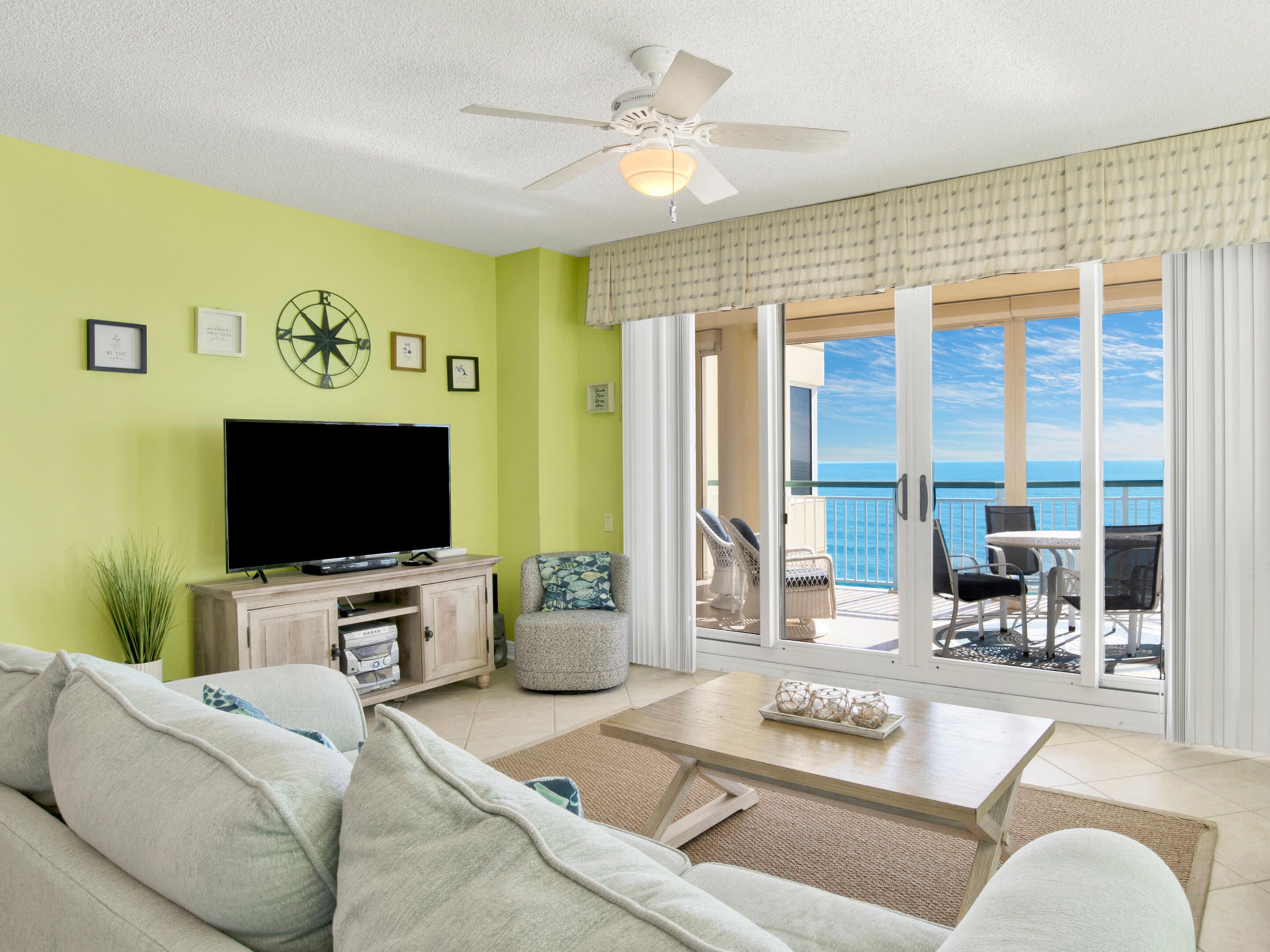 A living room with a flat-screen TV, a ceiling fan, and a large window overlooking the ocean.