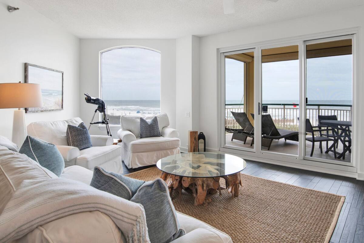 A spacious living room with a large window overlooking the beach, a ceiling fan, and a round glass coffee table.