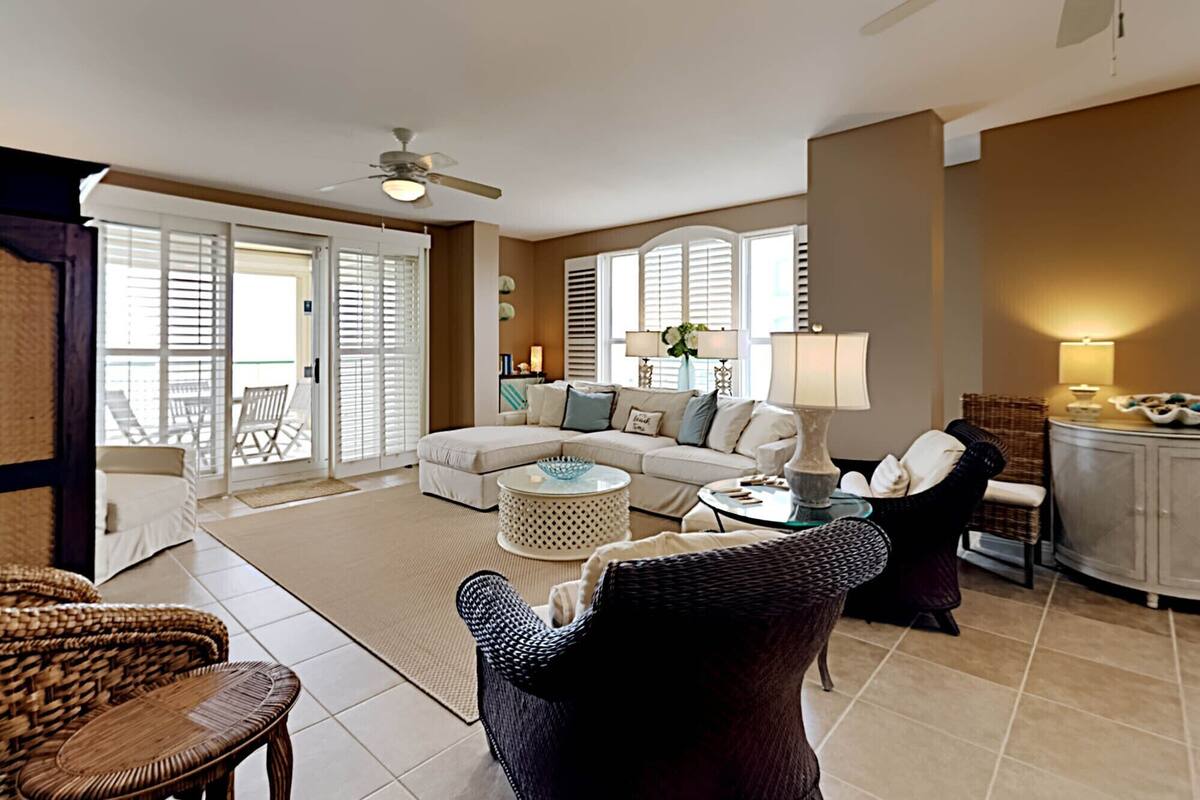 A spacious living room with a sofa, coffee table, and wicker chairs. There is a ceiling fan, a fireplace, and a view of the outdoors through large windows.