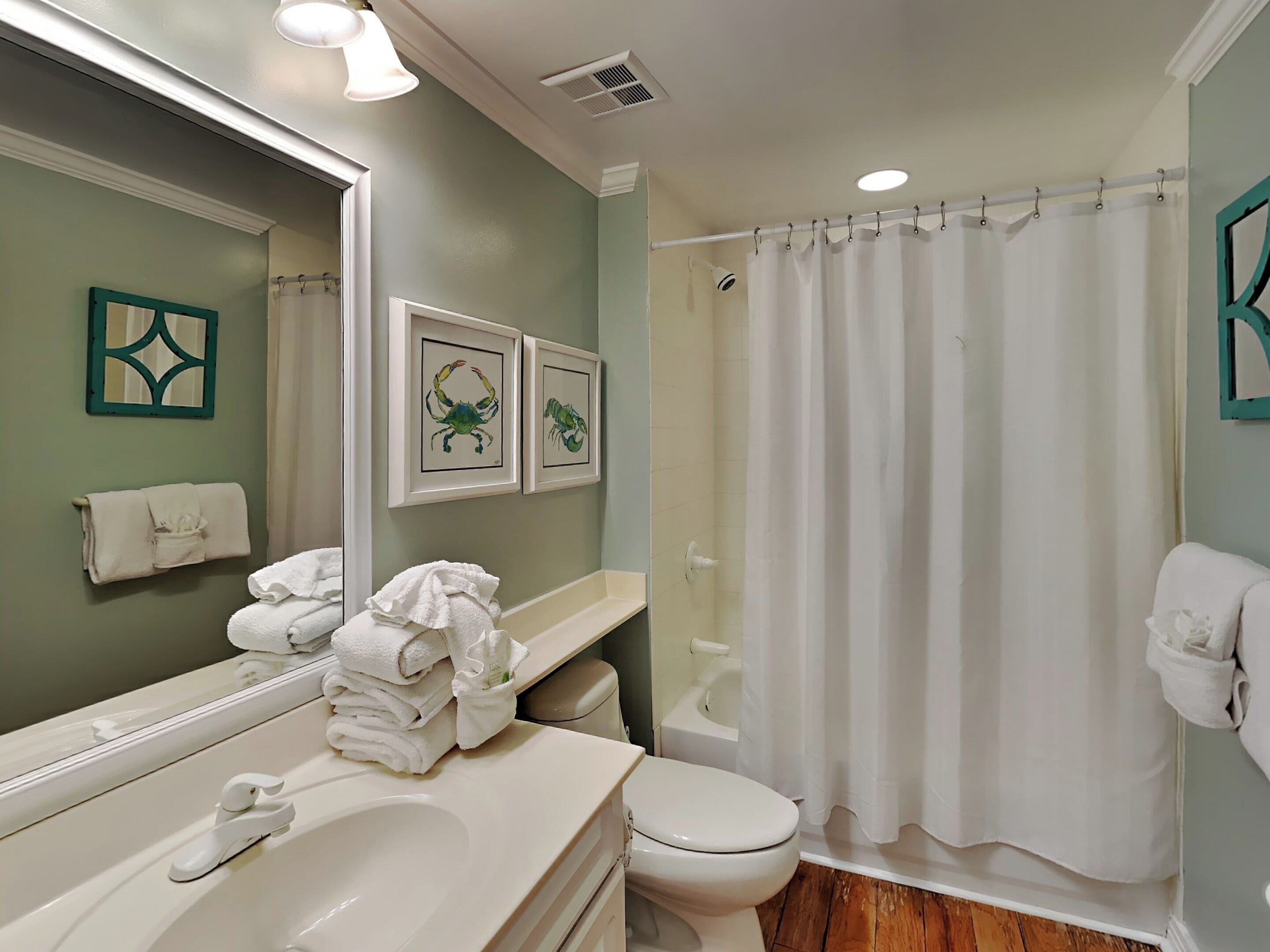 A bathroom with a sink, toilet, and shower curtain. There are towels stacked on the sink and a framed picture on the wall.