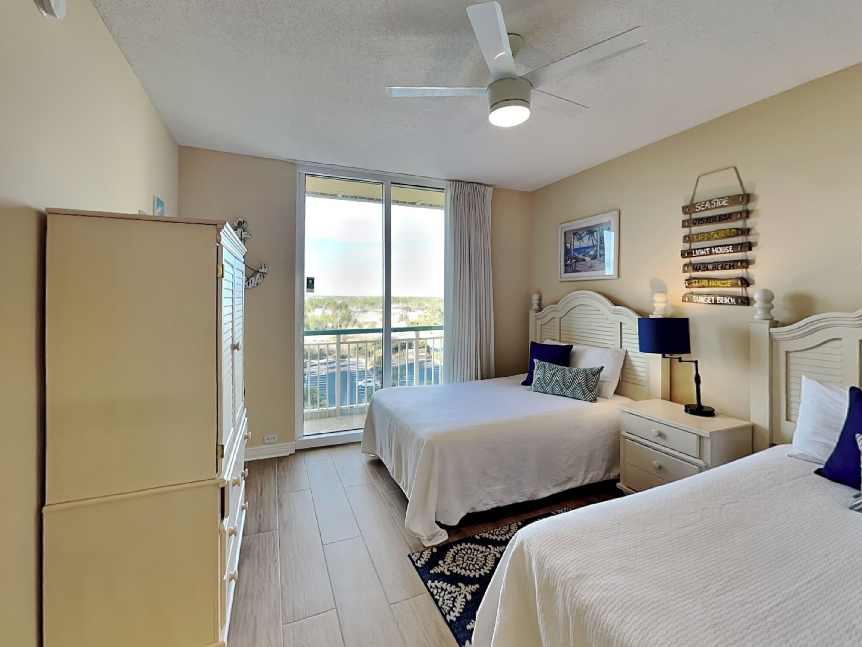 A hotel room with two beds, a ceiling fan, a large window with a view, a nightstand, and a wall-mounted sign with various names.