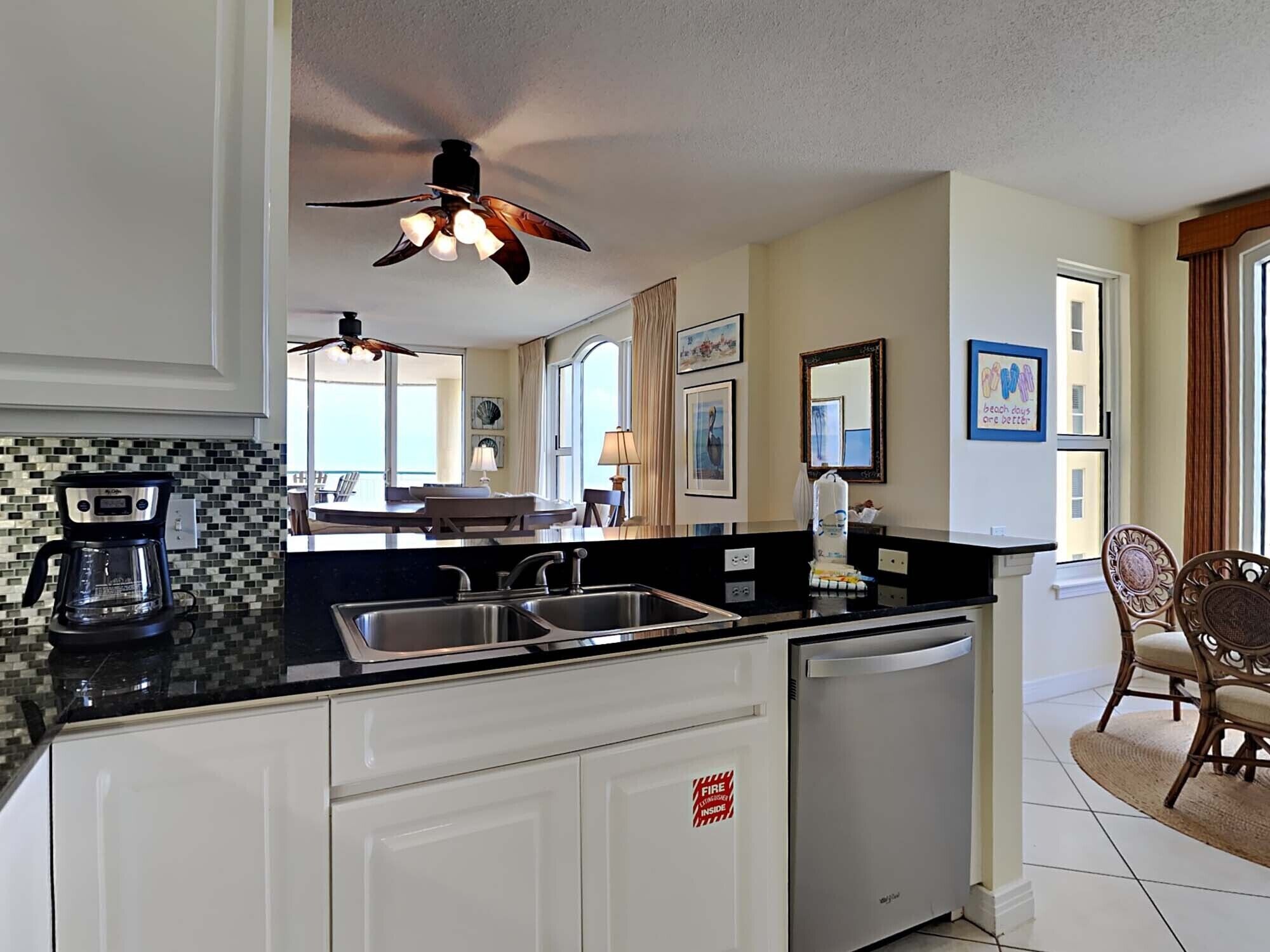 A modern kitchen with a black countertop, stainless steel sink, and a ceiling fan. There is a dining area with a table and chairs visible through the window.