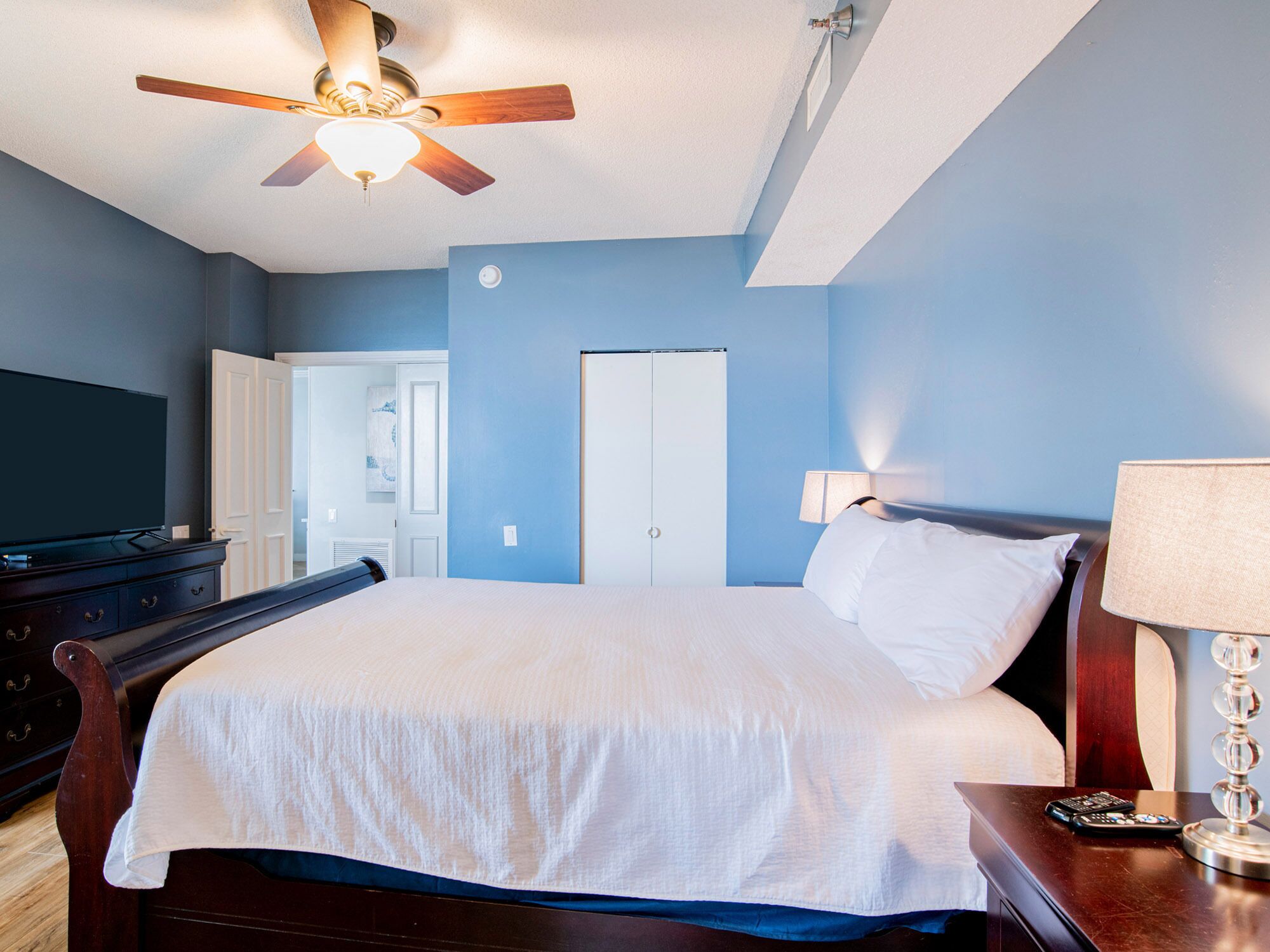 Une chambre d’hôtel équipée d’un lit, d’une télévision, d’un ventilateur de plafond, d’une table de chevet et d’une lampe.