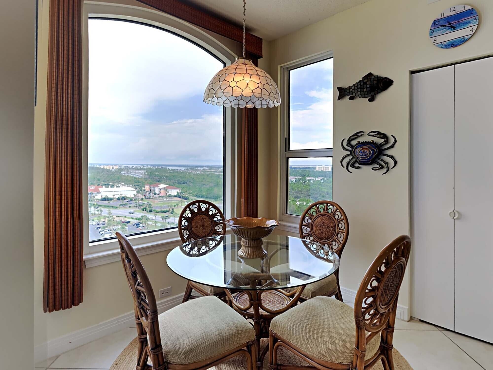 A dining area with a round glass table, four wicker chairs, a decorative hanging light, and a view of a cityscape through large windows.