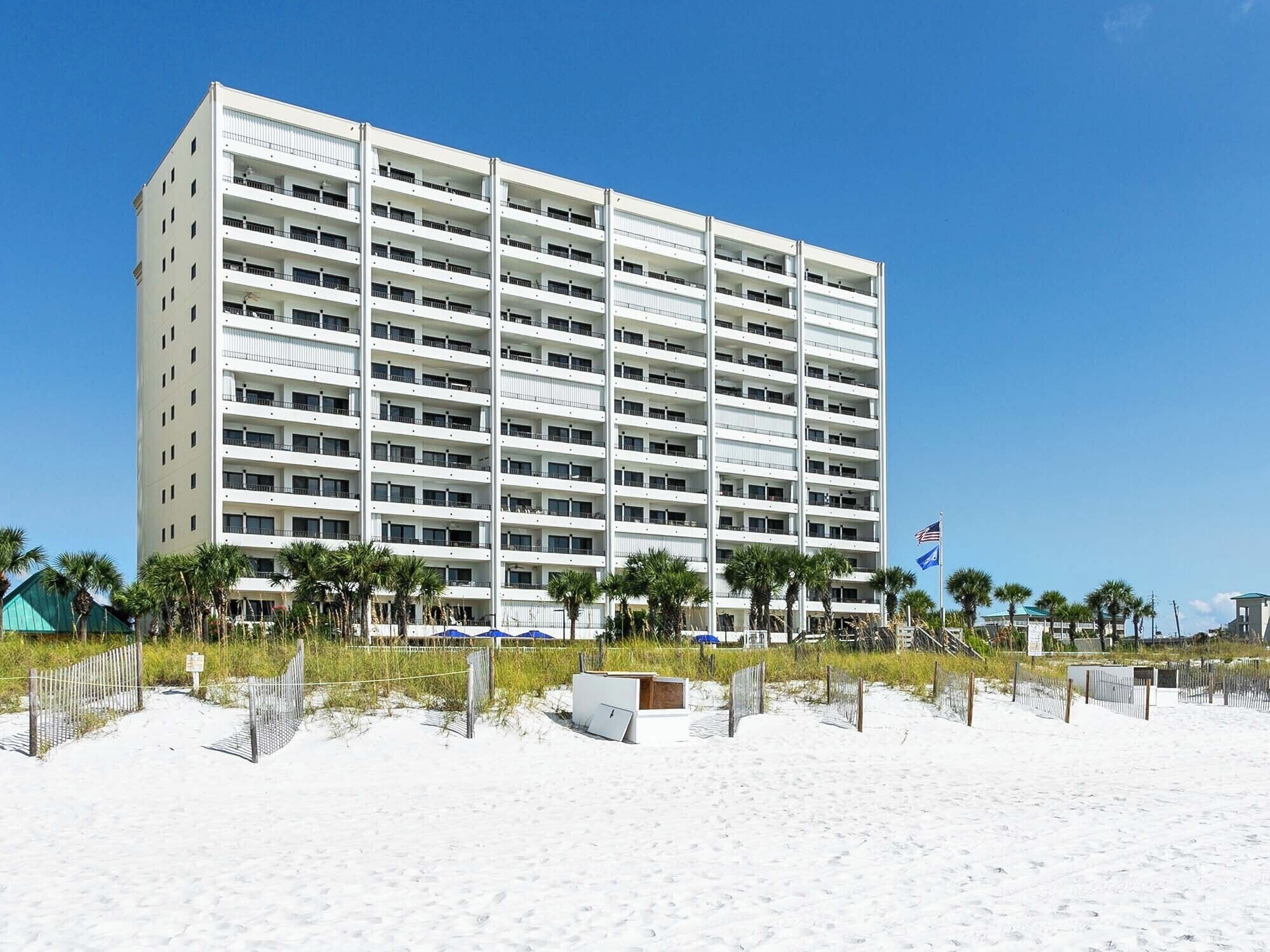 Un hôtel à plusieurs étages, doté de balcons, situé près d’une plage de sable bordée de palmiers.
