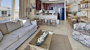 A modern living room with a sofa, a coffee table, and a patterned armchair. There is a kitchen area with a refrigerator, a dining table with chairs, and a bar area with stools. - Beach Club by Southern Vacation Rentals (Pensacola Beach)