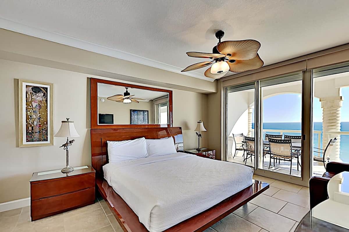 Un dormitorio amplio con una cama grande, ventilador de techo, balcón con mobiliario exterior y vista al océano.