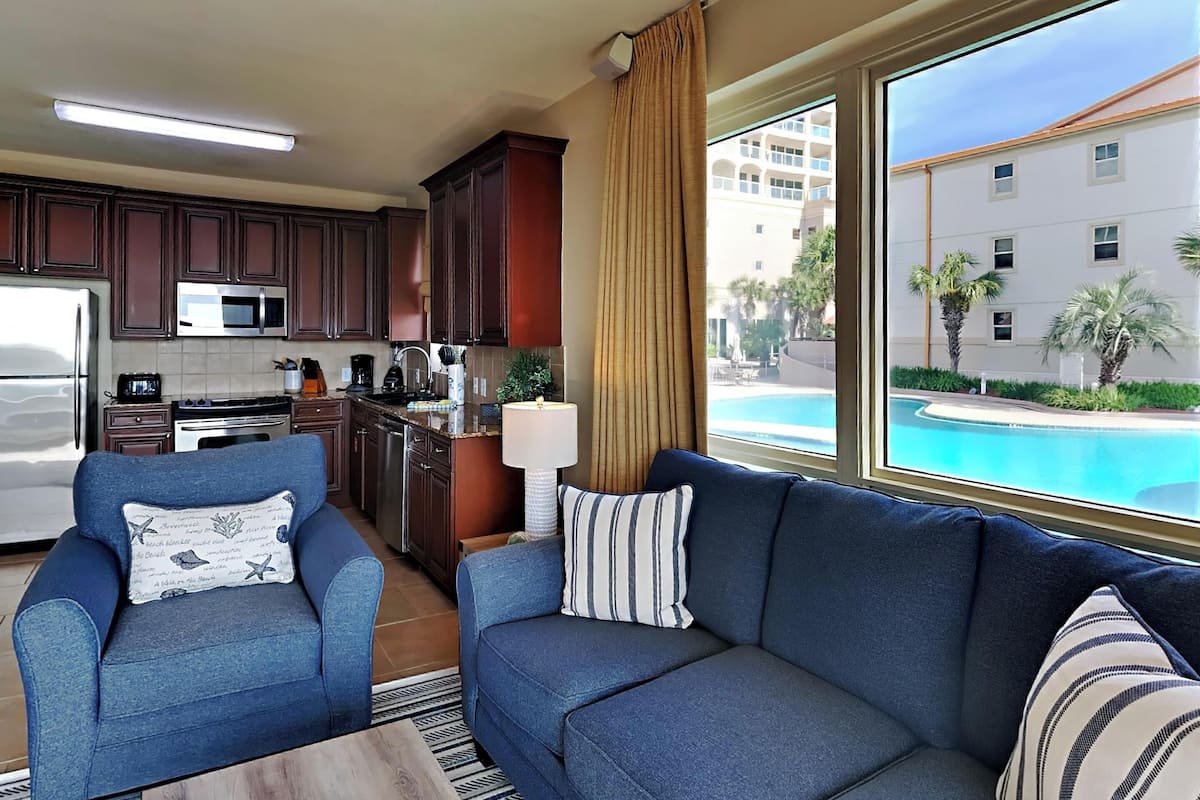 Una sala moderna con sofá y sillón, una cocina con armarios de madera oscura, microondas y vistas a una piscina y edificios.