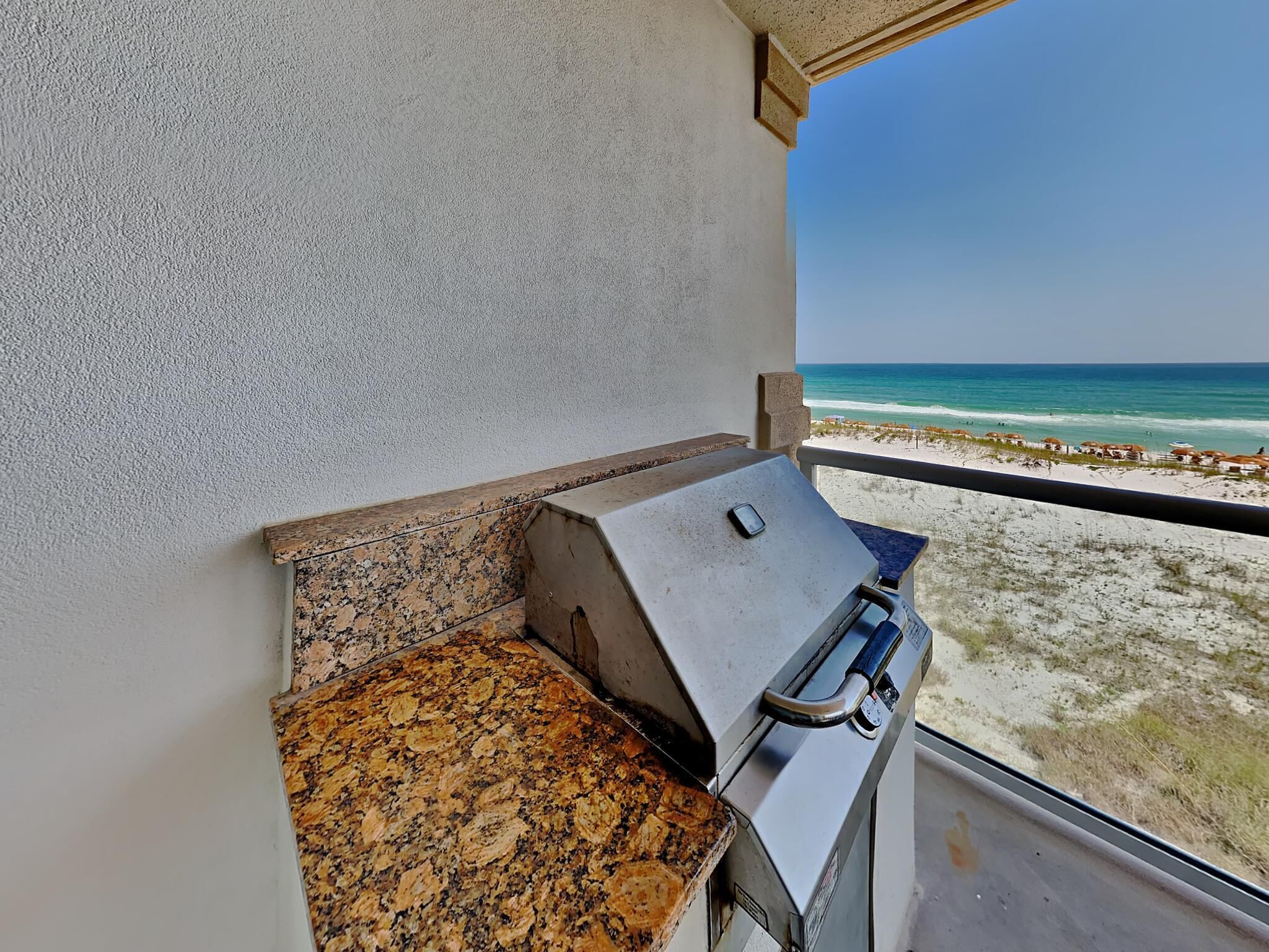 A balcony with a grill, a view of the beach, and a clear sky.
