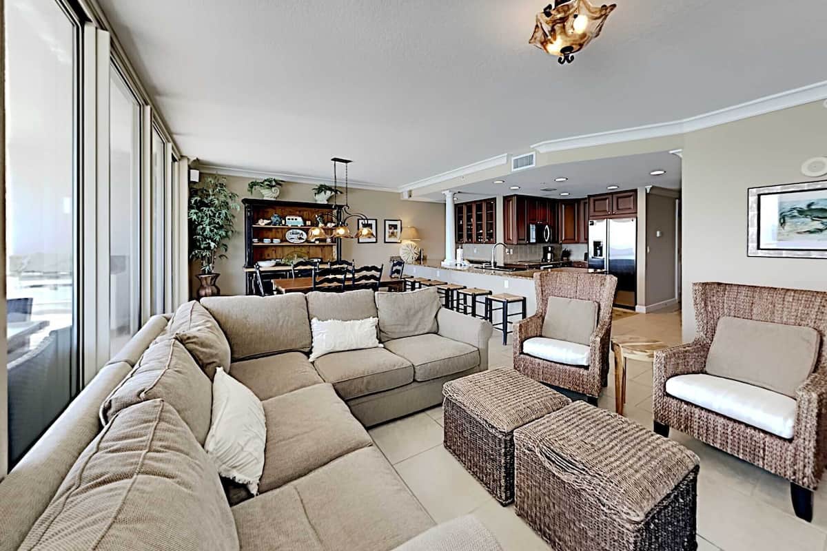 Amplia sala de estar con un sofá grande, dos sillones y una mesa de centro. Se observa una zona de cocina con armarios de madera oscura y una mesa de comedor.