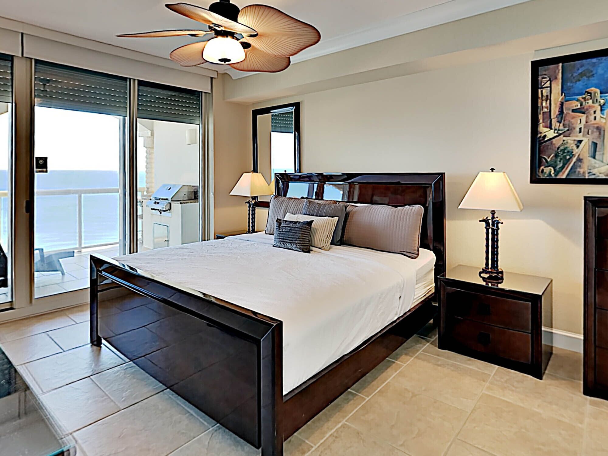 A bedroom with a large bed, two bedside tables, a ceiling fan, and a view of the sea through a sliding door.