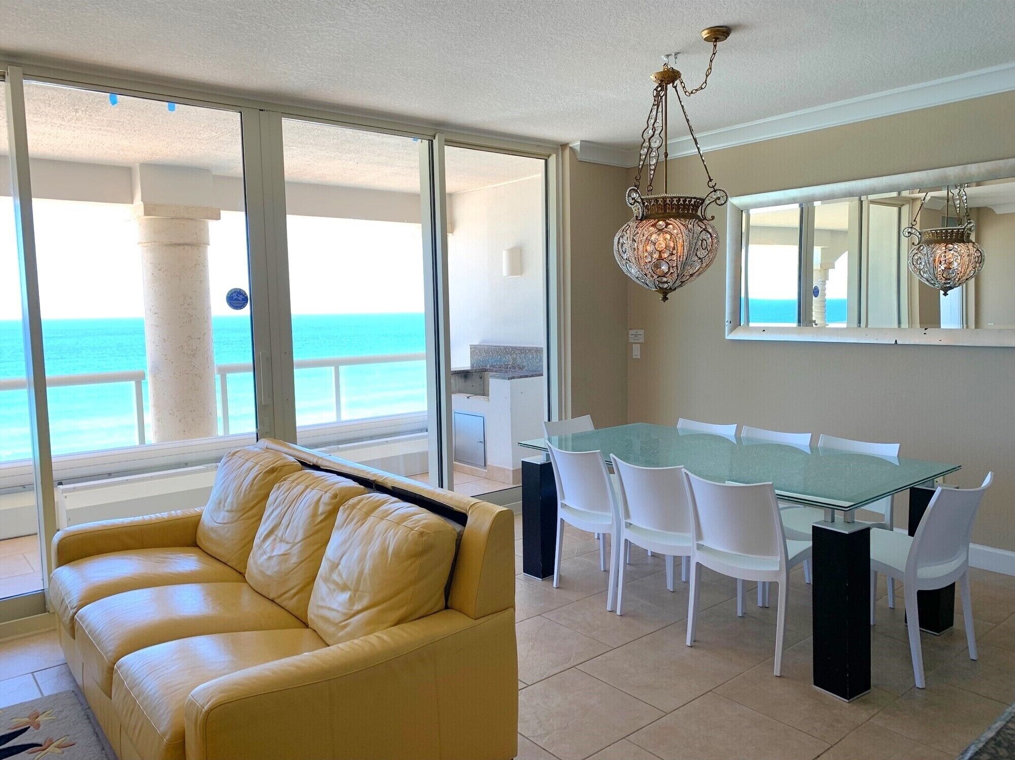 A modern living room with a glass table and white chairs, a yellow leather sofa, and a view of the ocean.